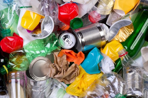A close-up image showing a pile of mixed waste materials, predominantly plastic bottles in transparent, green, and yellow colors, with some crumpled paper and metallic food cans with pull tabs. The plastic bottles vary in size and shape, with some caps attached and others loose. The crumpled paper is brown and appears to be discarded packaging. The metallic cans have smooth, shiny surfaces with visible ridges and pull tab openings. The waste is scattered across a dark background, likely a surface used for rubbish collection or disposal, illustrating typical household trash that a rubbish removal service such as Garden Clearance Kingston might handle within the Kingston area. The image emphasizes the diversity of recyclable and non-recyclable materials commonly collected during garden and rubbish clearance tasks in the local community.
