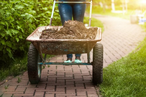 A person pushing a wheelbarrow filled with dark, moist soil along a paved garden pathway. The wheelbarrow has a metal frame, black rubber handles, and sturdy black wheels. The soil is slightly mounded, appearing rich and granular. The garden pathway is made of red-brick pavers and is bordered on the left by green foliage, including leafy bushes, and on the right by a well-maintained grassy lawn. In the background, there is a bright, sunlit area with additional greenery and garden elements, indicating a peaceful outdoor setting. The scene suggests a gardening or rubbish removal activity associated with garden clearance or general waste management, typical of services offered by Garden Clearance Kingston in the Kingston area.