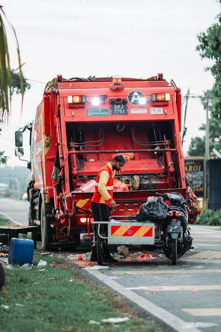 A roadside pile of waste consisting of black plastic trash bags, some partially torn open revealing mixed rubbish inside, along with discarded items such as a yellow plastic container, an old, dirty b