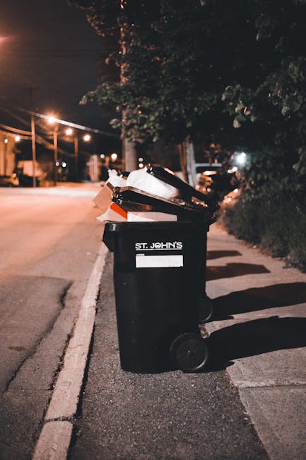 A black wheelie bin labeled 'St. John's' positioned on the edge of a quiet street at night, with its lid open and filled with assorted rubbish including white and brown paper bags and cardboard. The b