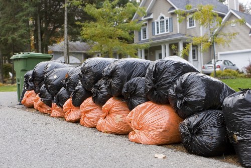 A row of tightly packed black and orange plastic rubbish bags filled with waste materials, lined up along the edge of a residential street pavement in a suburban neighbourhood. The black bags dominate the foreground, with their shiny, crinkled surfaces reflecting light, while several orange bags are positioned at the bottom of the row, possibly containing garden debris or general refuse. Behind the bags, a green wheelie bin is visible, suggesting disposal collection is imminent. The background features a quiet street with a mix of parked cars, a driveway, and a detached house with light-colored exterior walls, multiple windows, and a sloped roof, surrounded by trees with green foliage. The overall scene is captured during daylight, with natural lighting highlighting the textures of the bags, the pavement, and the nearby greenery, indicative of typical rubbish clearance activity near residential properties within the Kingston area, where Garden Clearance Kingston offers waste removal services.